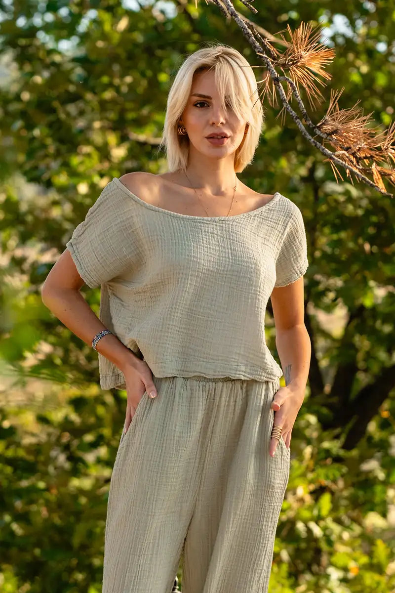 Woman wearing a light green gauze top and pants standing in a natural setting with trees.