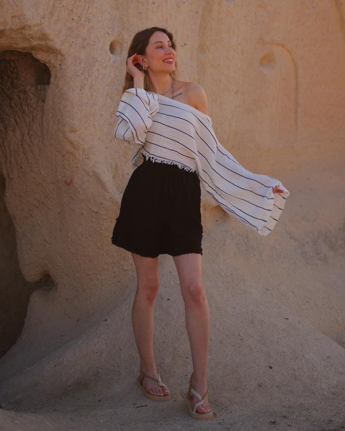 A woman in a striped top and cotton gauze shorts stands confidently in front of a large rock formation.