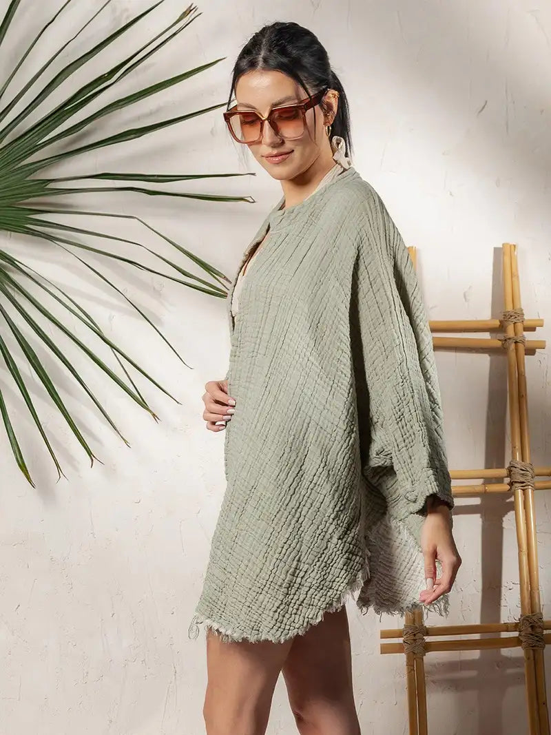 Woman wearing a green textured cotton long sleeve cardigan with sunglasses, standing against a white wall with a plant and bamboo rack.