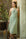 Woman in a light green halter neck dress standing next to a textured wall with a plant and suitcase in the background.