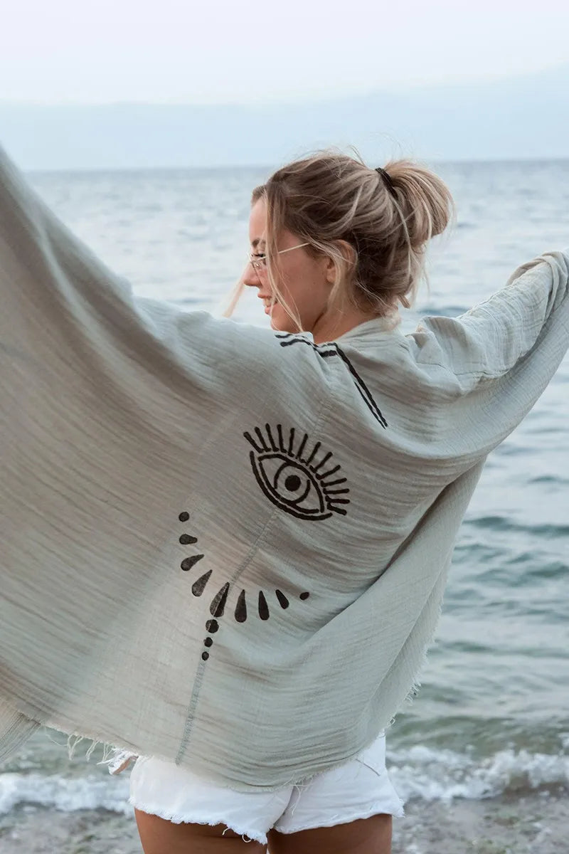 Woman standing by the ocean wearing a green double gauze cotton cover-up with an evil eye design.