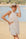 Woman in a beige outfit and hat standing on a beach with water and sky in the background