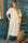 Woman in a shirt dress standing against a rustic wooden wall with a woven bag.