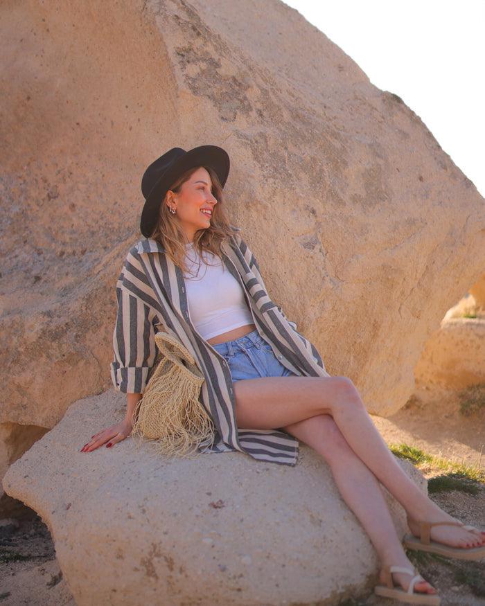  A woman wearing a striped midi shirt and hat sits on a rock, enjoying the outdoors.