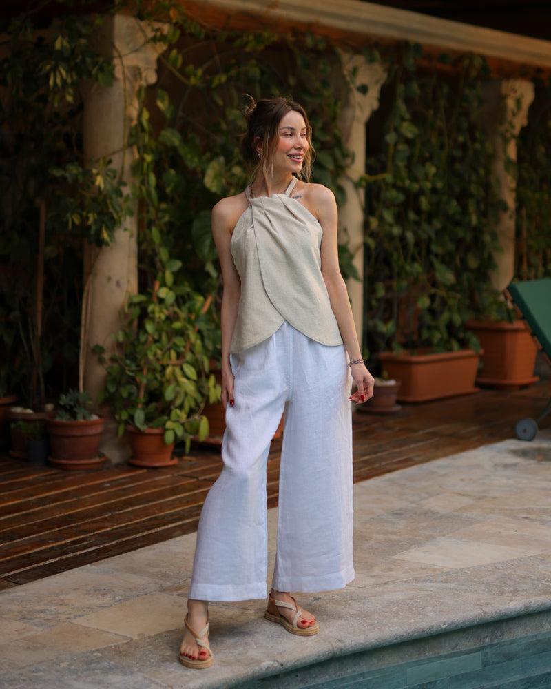 A woman wearing a beige linen sleeveless halter top and pants stands next to a pool, looking relaxed and stylish.
