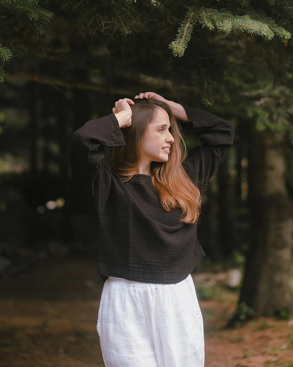 Woman in a black muslin sweater and white pants standing in a forest