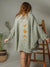 Person wearing a green crinkle cotton textured dress with gold moon phases design, standing in a minimalistic room.