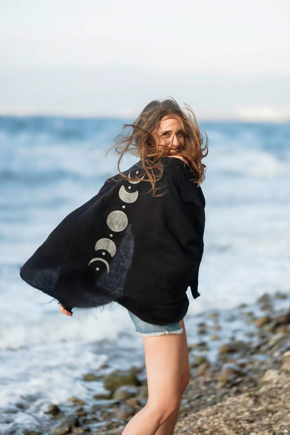 Woman standing on a beach wearing a black cardigan with celestial moon phases designs.