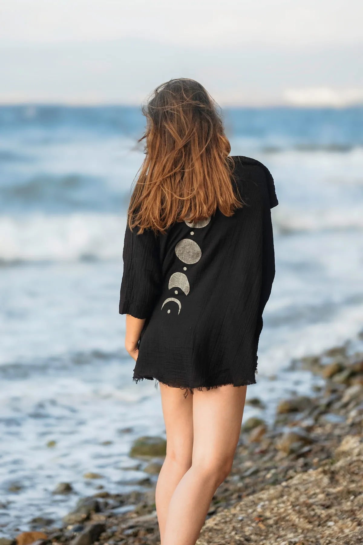 Person wearing a black Crinkle Cotton  Jacket with Silver Moon design standing on a rocky beach.