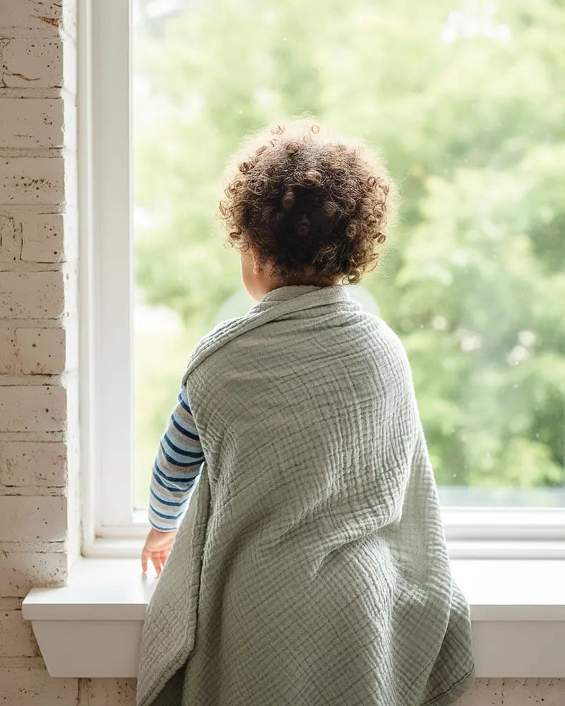 Child wrapped in a muslin blanket looking out a window with a view of greenery