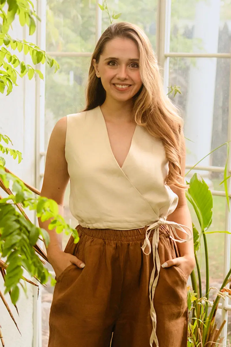 Woman in a beige tie front vest top and brown pants standing among plants indoors