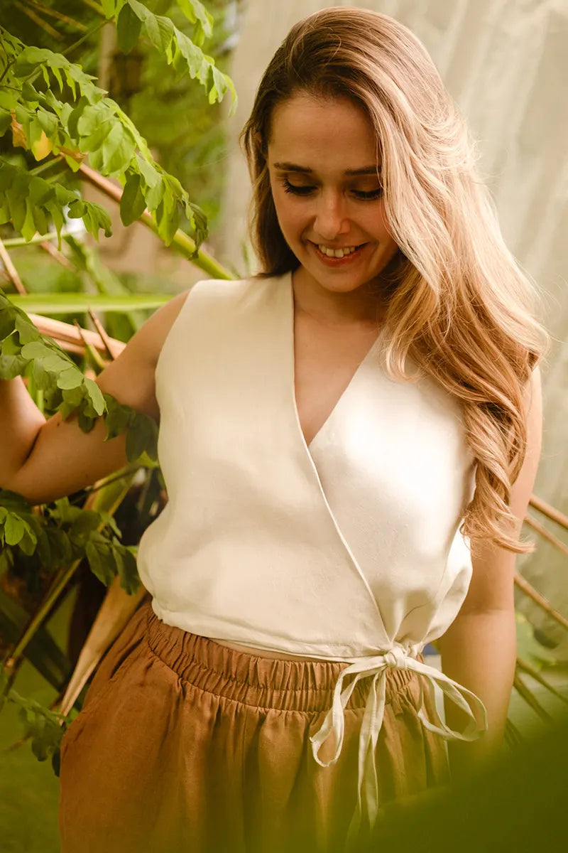 Woman in a beige linen sleeveless vest top and brown pants standing among green foliage.
