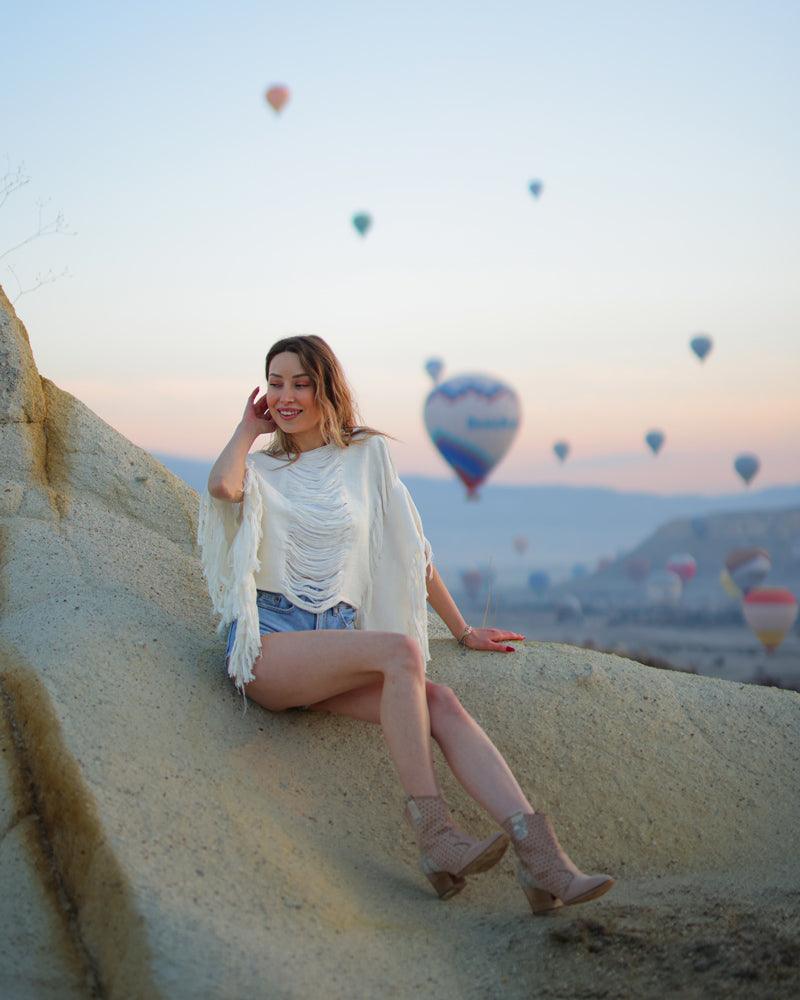 A woman in a fringe top sits on a rock, with colorful hot air balloons floating in the background.