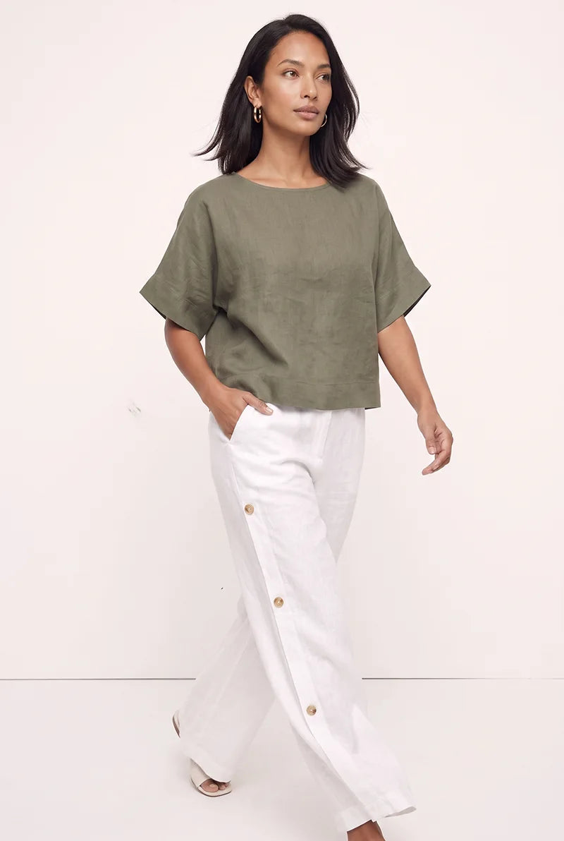 A woman modeling a short-sleeve olive green top and white wide-leg pants with gold-tone side buttons, standing against a plain off-white studio background.
