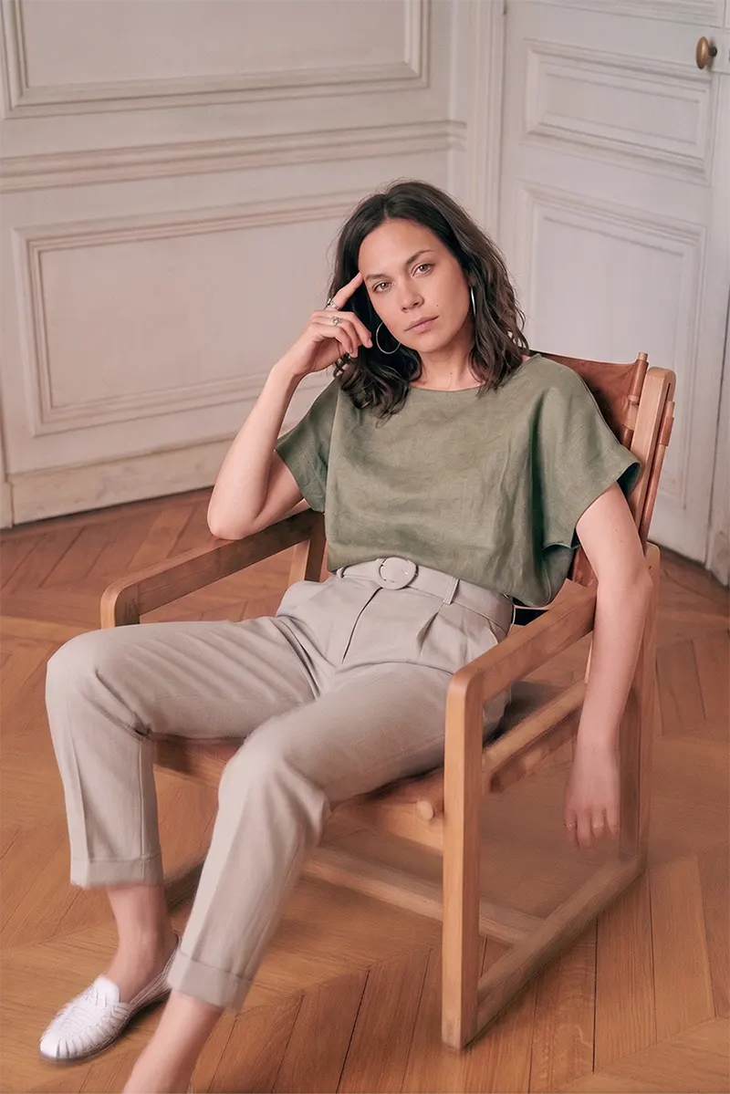 Woman seated in an armchair wearing the olive green linen blouse top with beige trousers