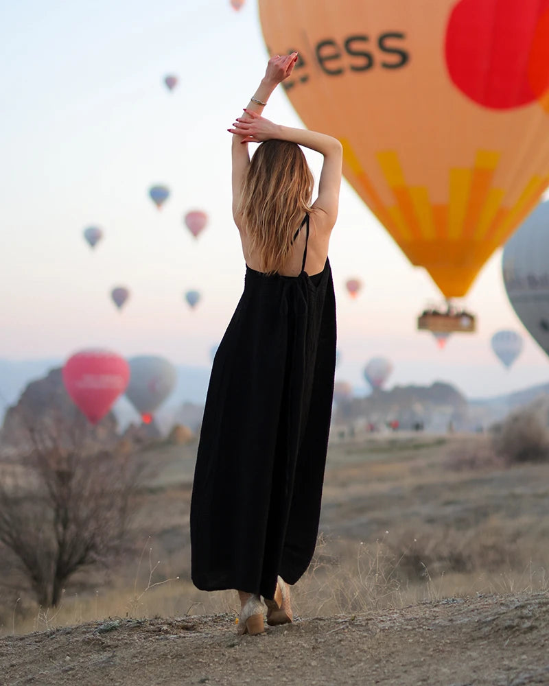 Woman in a open back black muslin dress standing with arms raised against a backdrop of hot air balloons.