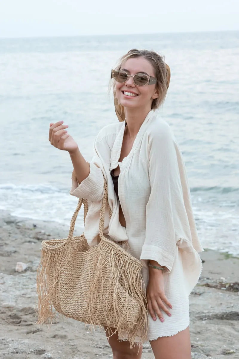 Woman on a beach wearing sunglasses and a cream muslin cover up with a beige bag.