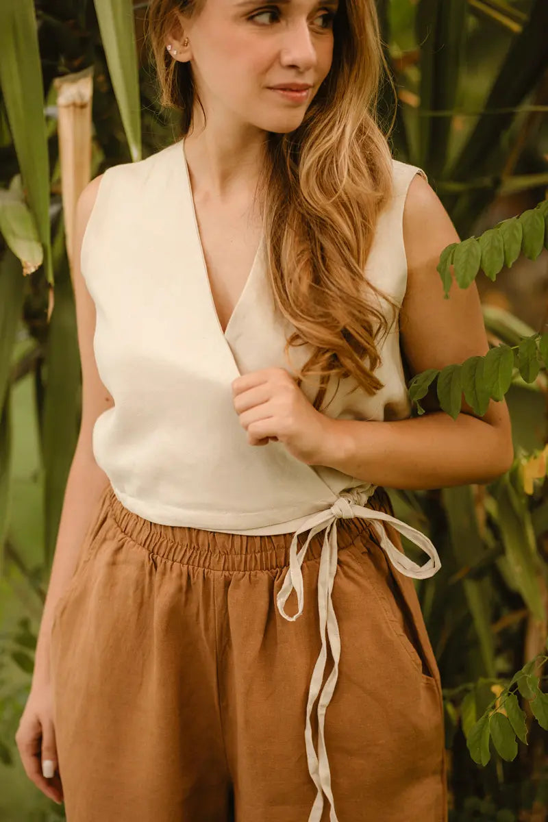 Woman wearing a beige sleeveless tie front vest top and brown pants with a green leafy background