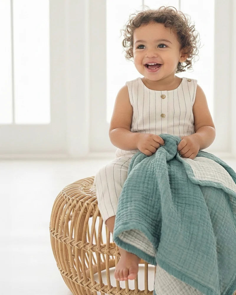 Child sitting on a wicker stool holding a muslin blanket in a bright room.