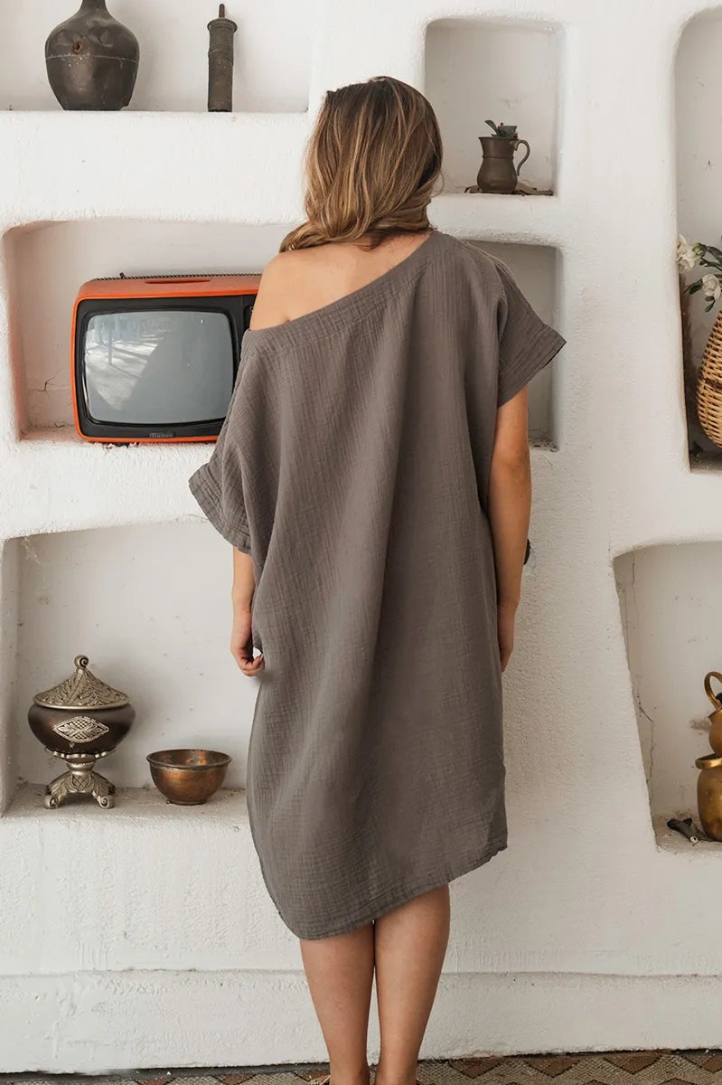 Woman wearing a gray off-shoulder flowy tunic dress standing in a room with white walls and decorative items.