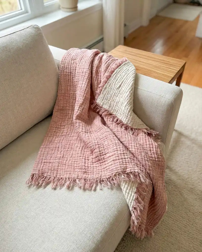 Pink cotton gauze blanket draped over a beige sofa in a cozy living room.