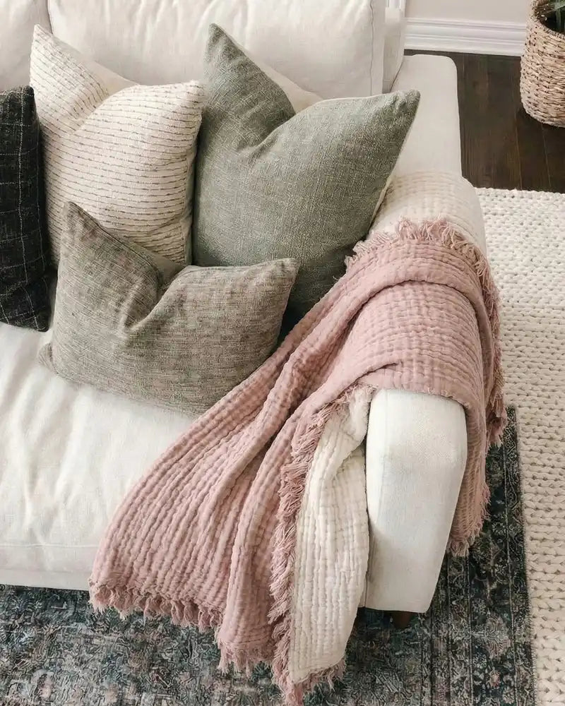 White sofa with textured pillows and a pink gauze blanket on a dark wooden floor.