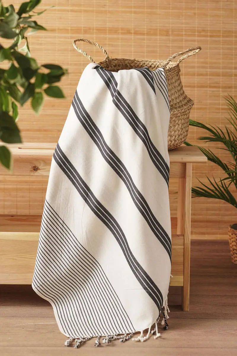 White turkish towel with black stripes and tassels on a wooden surface with plants in the background