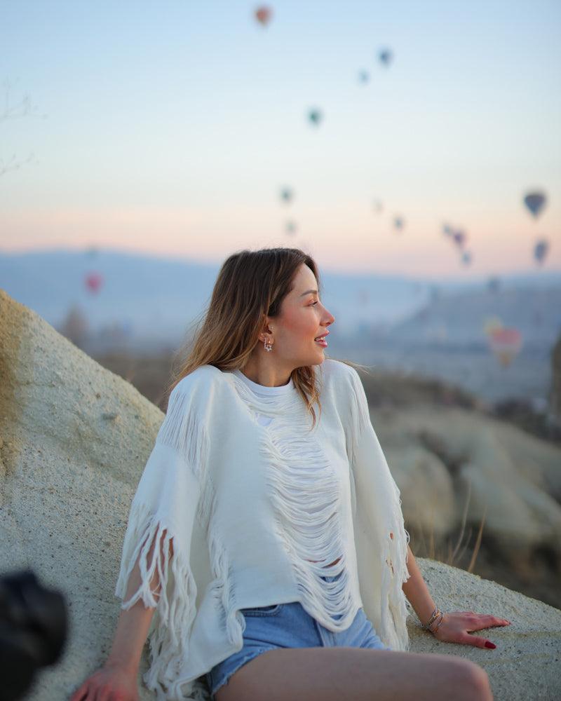 A woman sits on a rock with fringe blouse, gazing at colorful hot air balloons floating in the sky behind her.