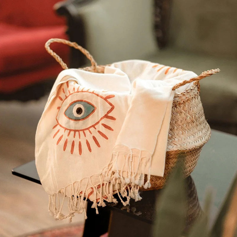 Decorative eevil eye design turkish beach towel draped over a woven basket on a glass table.