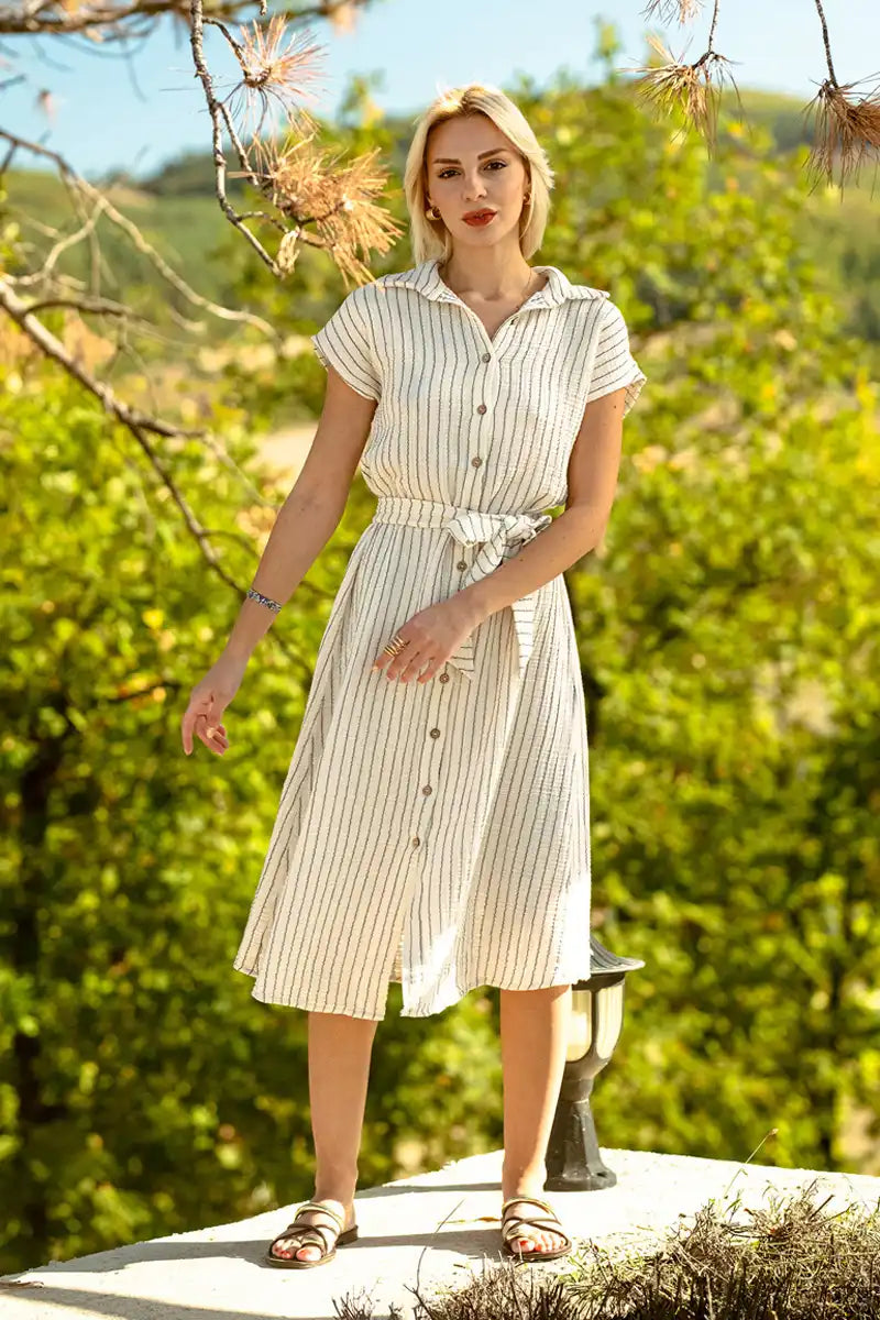 Woman in a short sleeve belted dress standing outdoors with greenery in the background