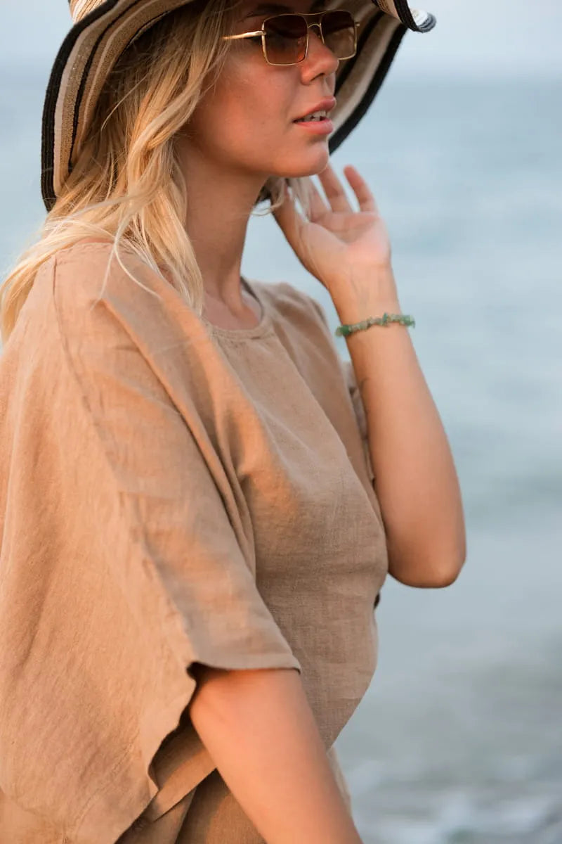 Woman wearing a brown linen blouse and hat with sunglasses, standing against a blurred natural background