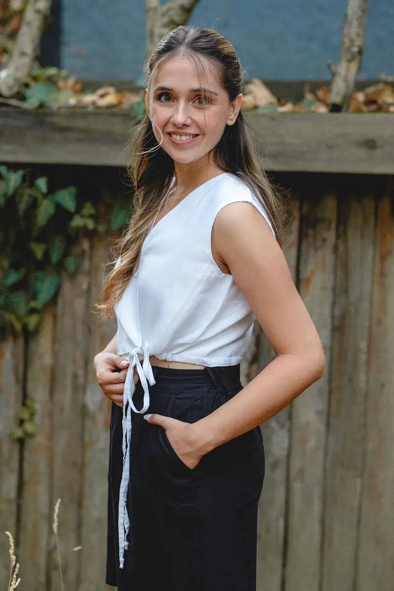 Woman wearing a white sleeveless linen vest top and black pants standing outdoors with a wooden fence and plants in the background.