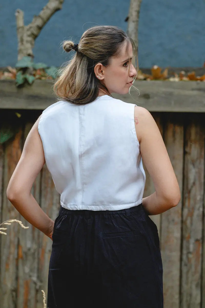 Woman wearing a white sleeveless  linen wrap top and black skirt standing outdoors with a wooden fence and trees in the background.