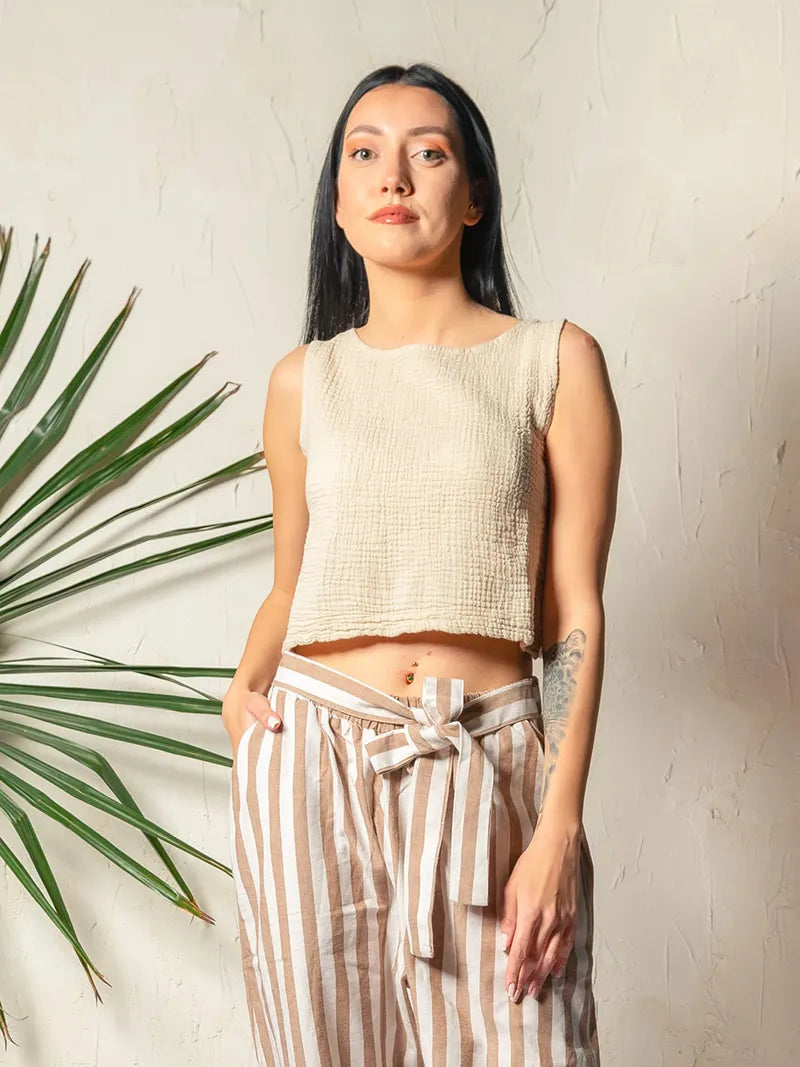 Woman wearing a beige sleeveless double gauze top and striped pants standing against a neutral background with a plant to the left.