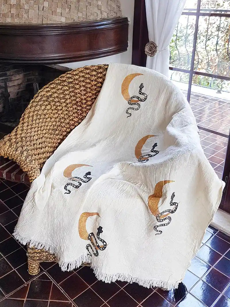 White blanket with celestial snake and moon designs draped over a chair in a room with a window and outdoor view.