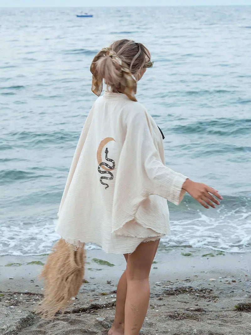 Woman standing on a beach wearing a beige kimono with a bohemian design, facing the ocean.