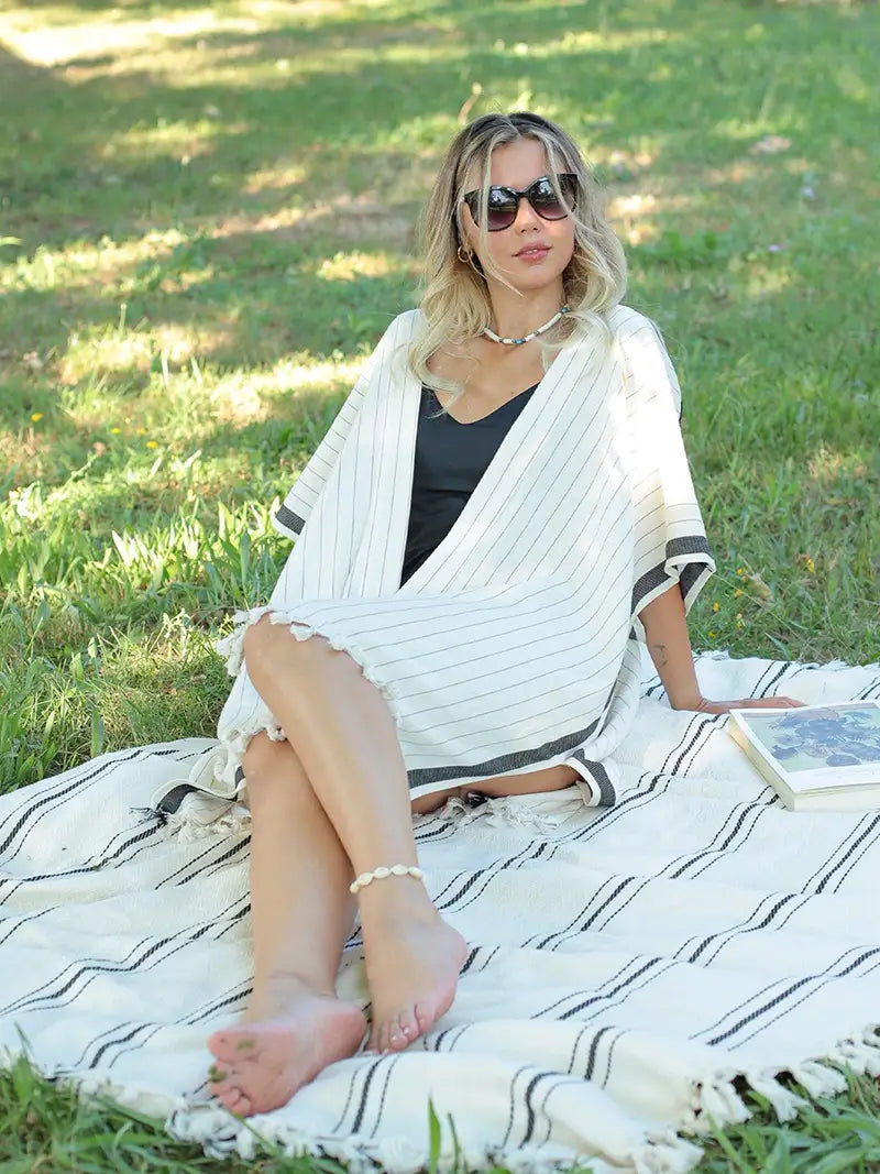 Woman sitting on a blanket in a grassy area wearing sunglasses and a black striped white cover-up.
