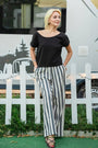 Woman in a black top and black and white striped linen pants by a white fence — linen-cotton blend, wide-leg cut