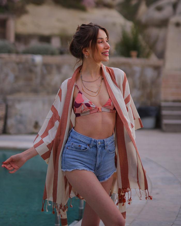 A person sitting on a lounger wearing a orange and white striped beach wrap dress with short denim shorts.