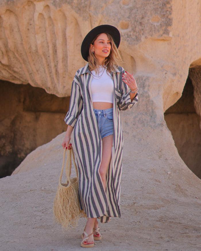 A woman standing in a casual pose wearing a striped button down maxi shirt dress with long sleeves, paired with a white top and blue shorts, accessorized with a wide-brimmed hat, a beaded necklace, and carrying a straw tote bag.