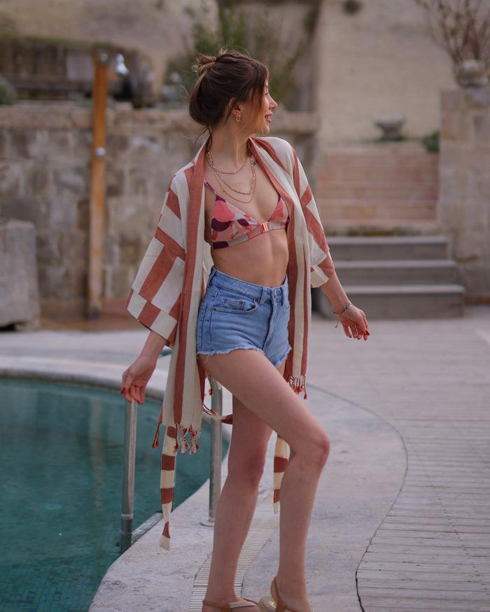  A woman in a bikini top and shorts stands by a pool, wearing a striped kimono.