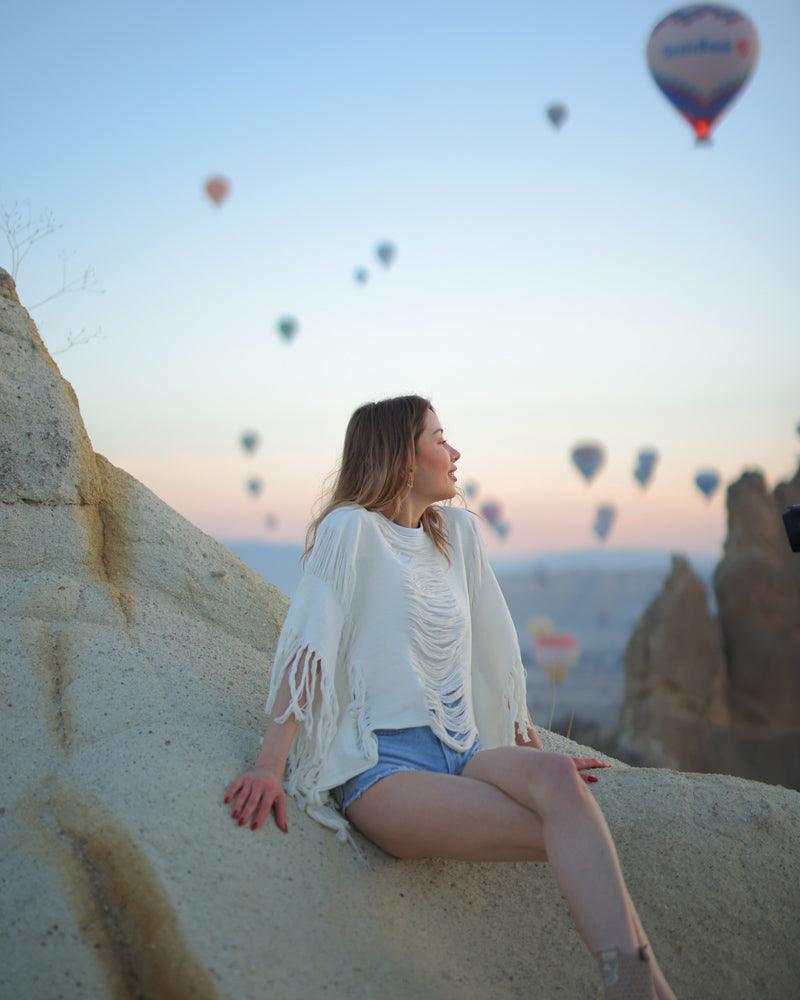 A woman in a Tulum kimono top sits on a rock, with colorful hot air balloons floating in the background.