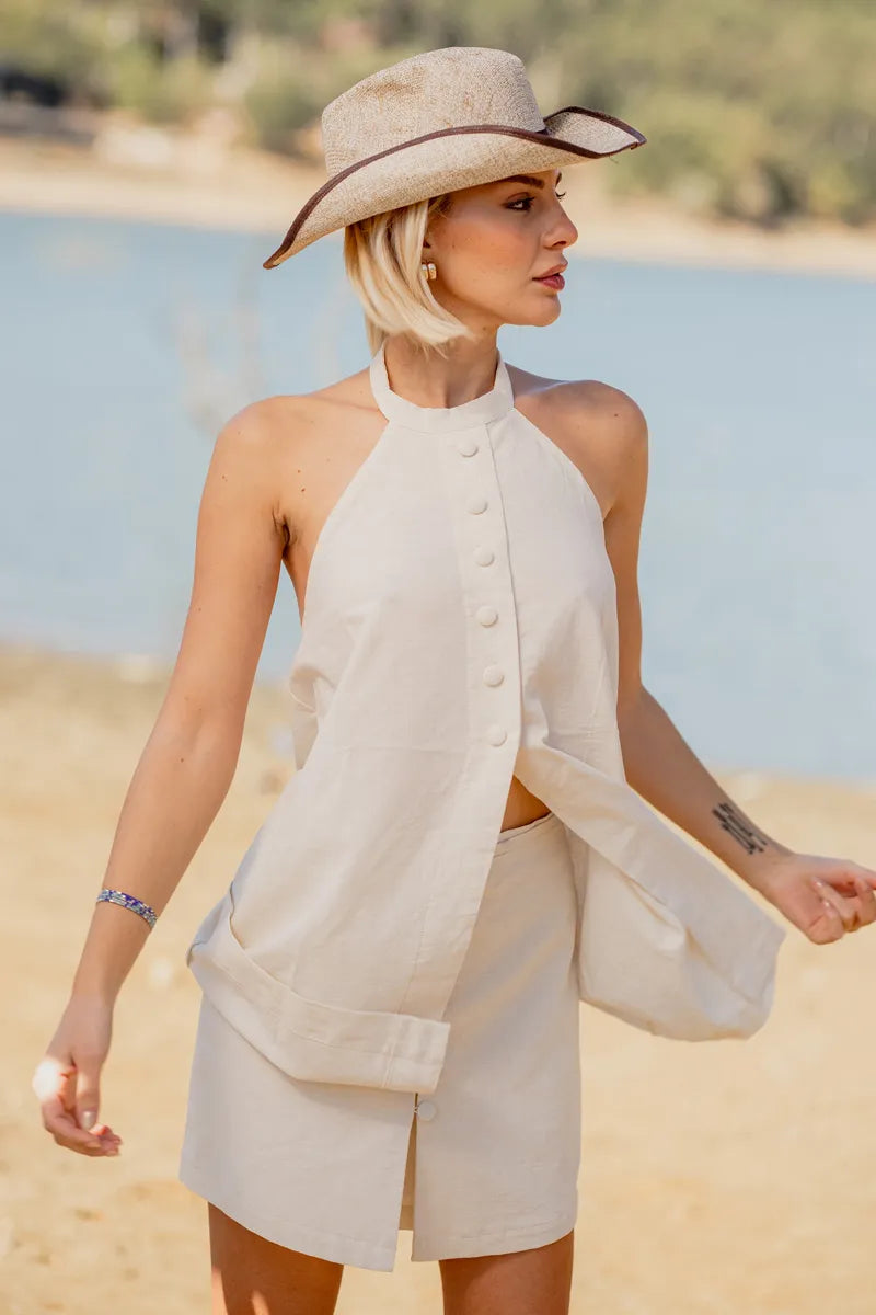 Woman in a beige vest and shorts outfit and hat standing on a beach with water and sky in the background