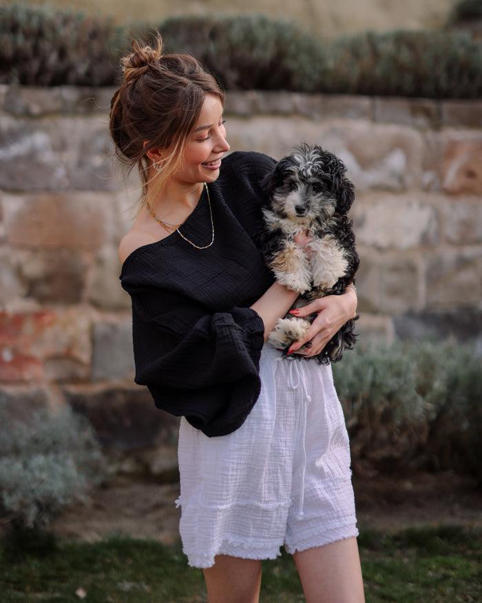 A woman in a black gauze top and white double gauze shorts holds a small dog, smiling at the camera.