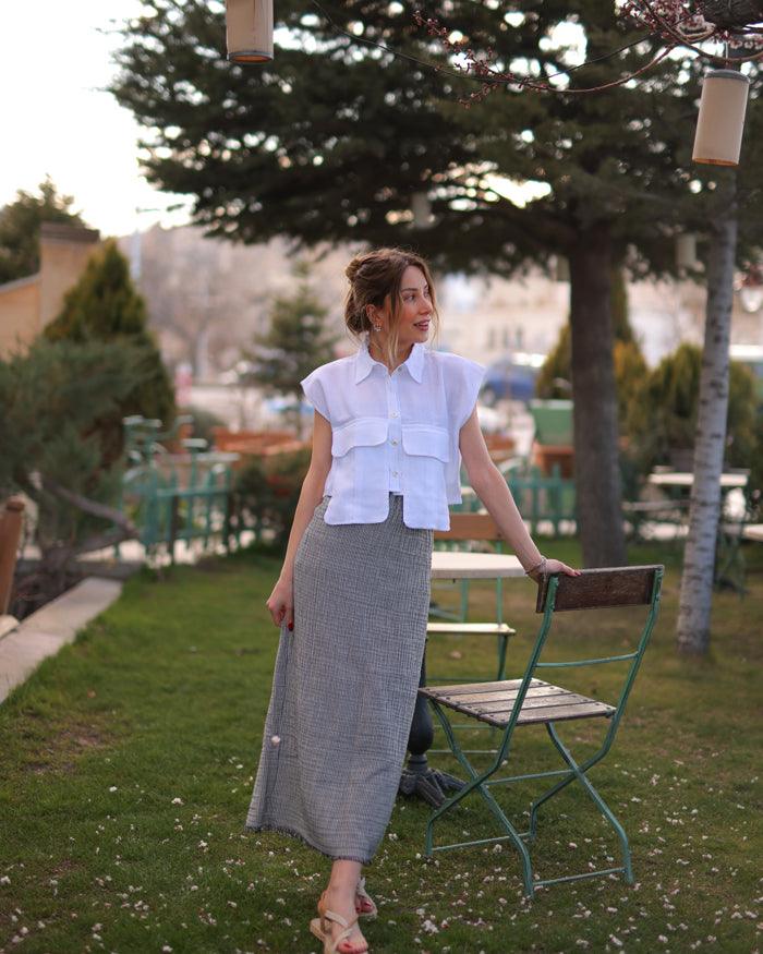 A woman in a white sleeveless collared shirt and grey skirt stands in the garden