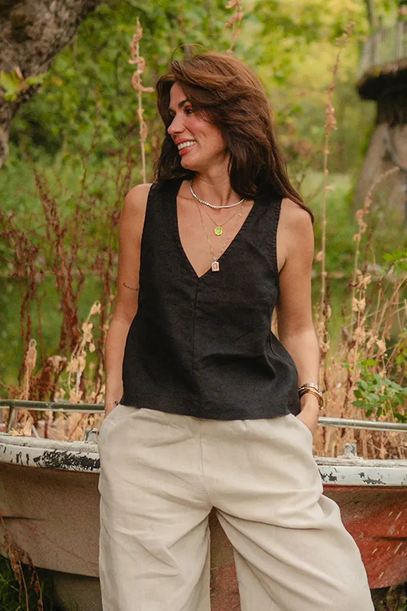 Woman wearing a black sleeveless v neck Linen Wide Strap Tank Top top and beige pants standing outdoors with greenery in the background